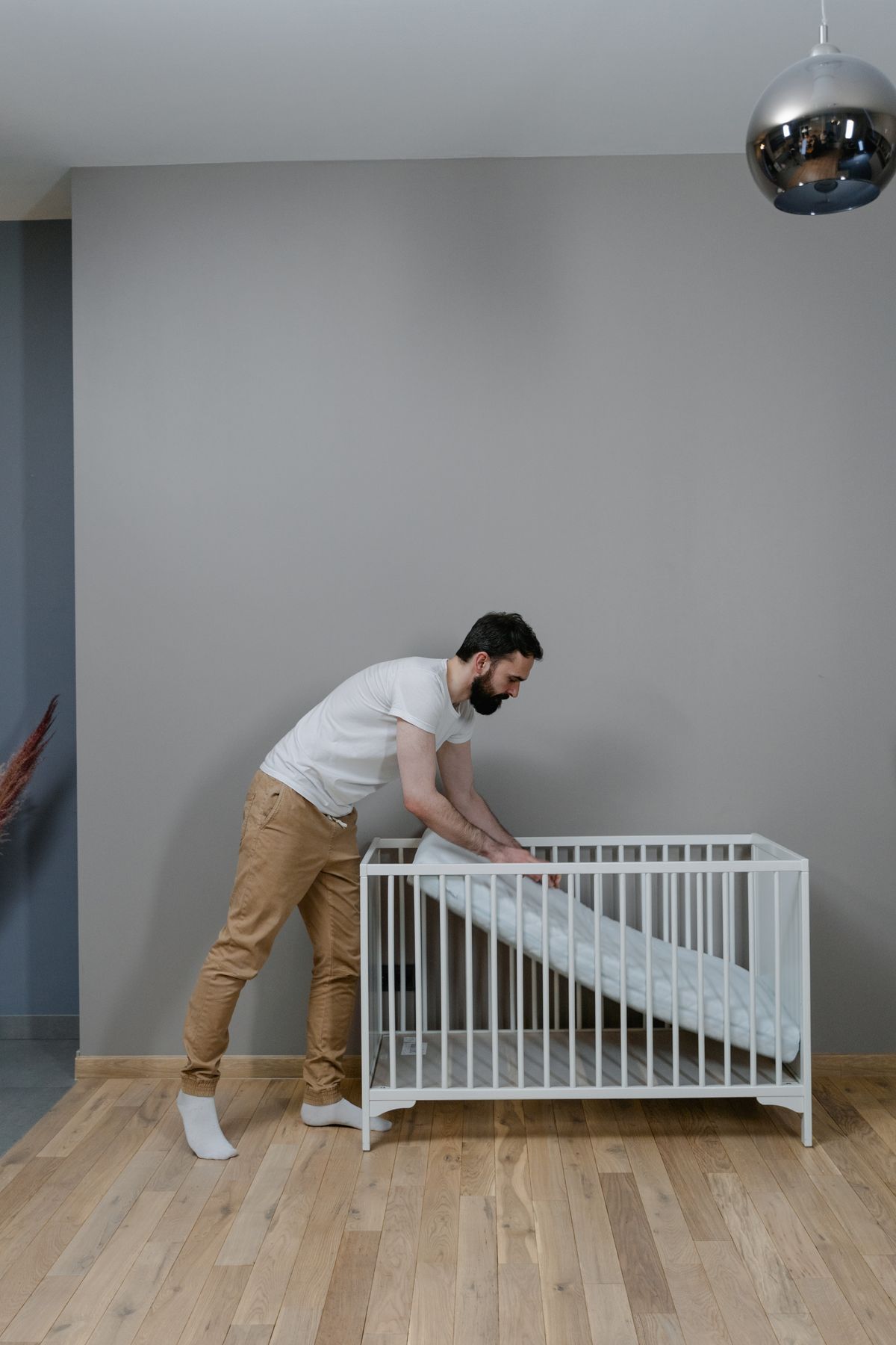 Young man adjusting a crib mattress in a modern, minimalist nursery with hardwood floors.