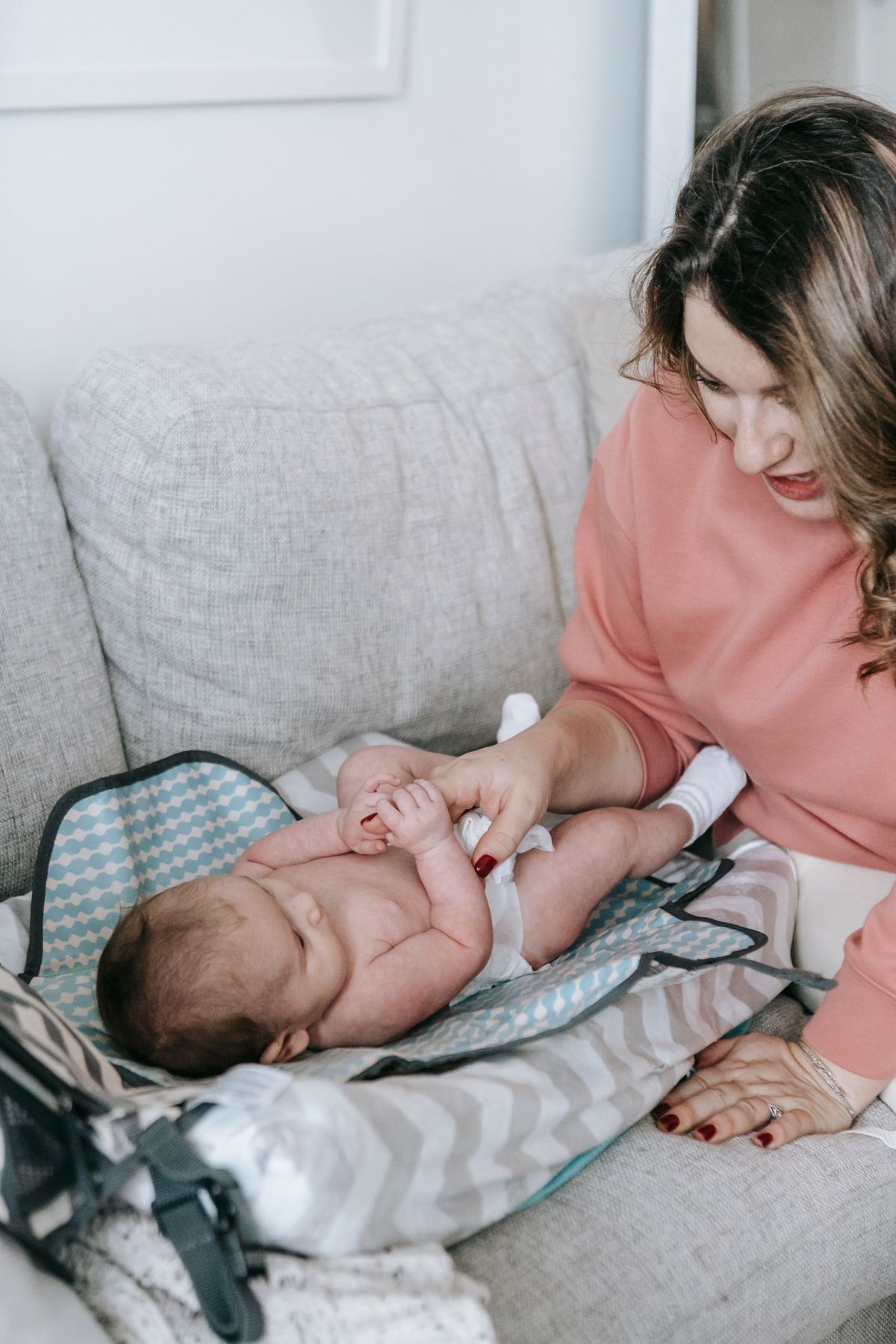 Caring mother in casual clothes playing with cute infant baby in diaper on comfortable sofa in light living room at home